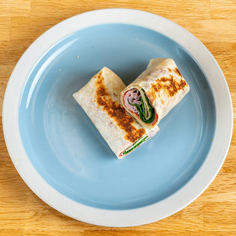 Two halves of a grilled ham and spinach wrap on a blue and white plate.