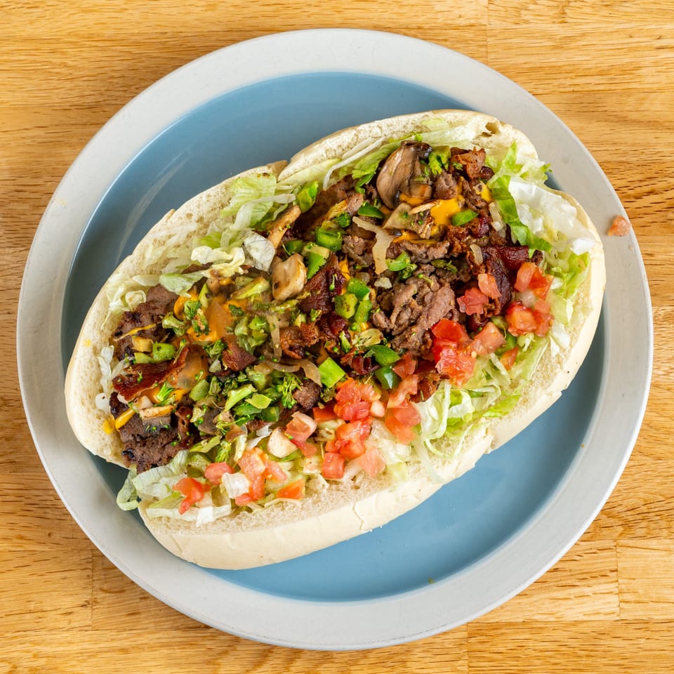 A large, open-faced steak and cheese sub with lettuce, tomatoes, mushrooms, and green peppers on a blue plate.