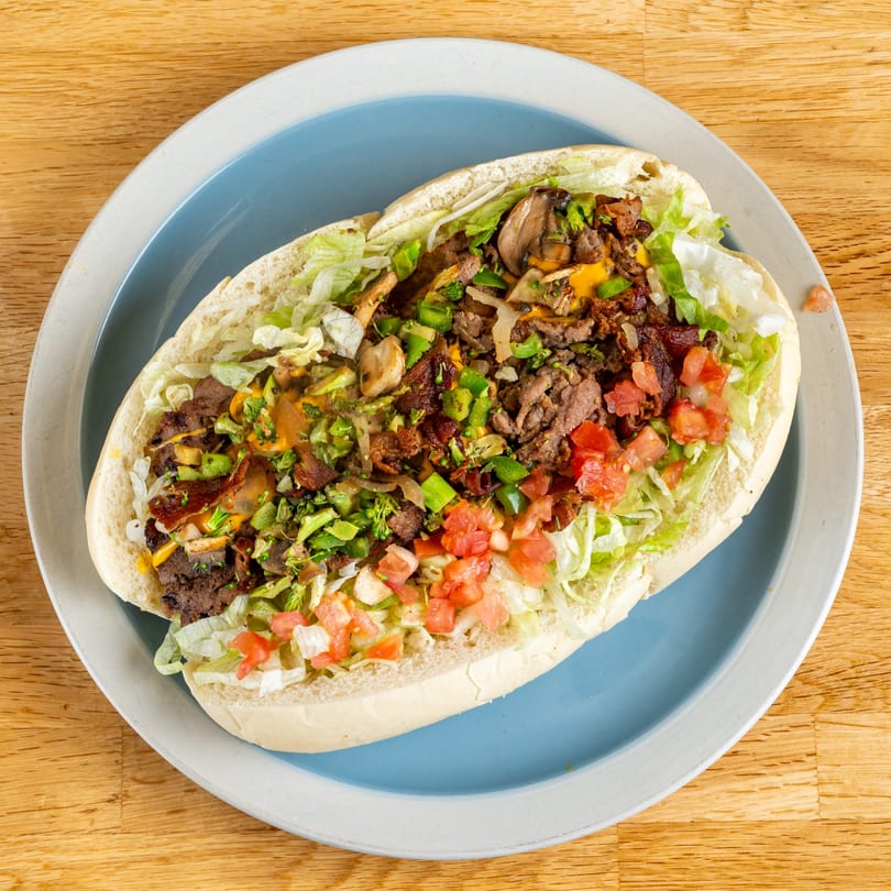 A large, open-faced steak and cheese sub with lettuce, tomatoes, mushrooms, and green peppers on a blue plate.