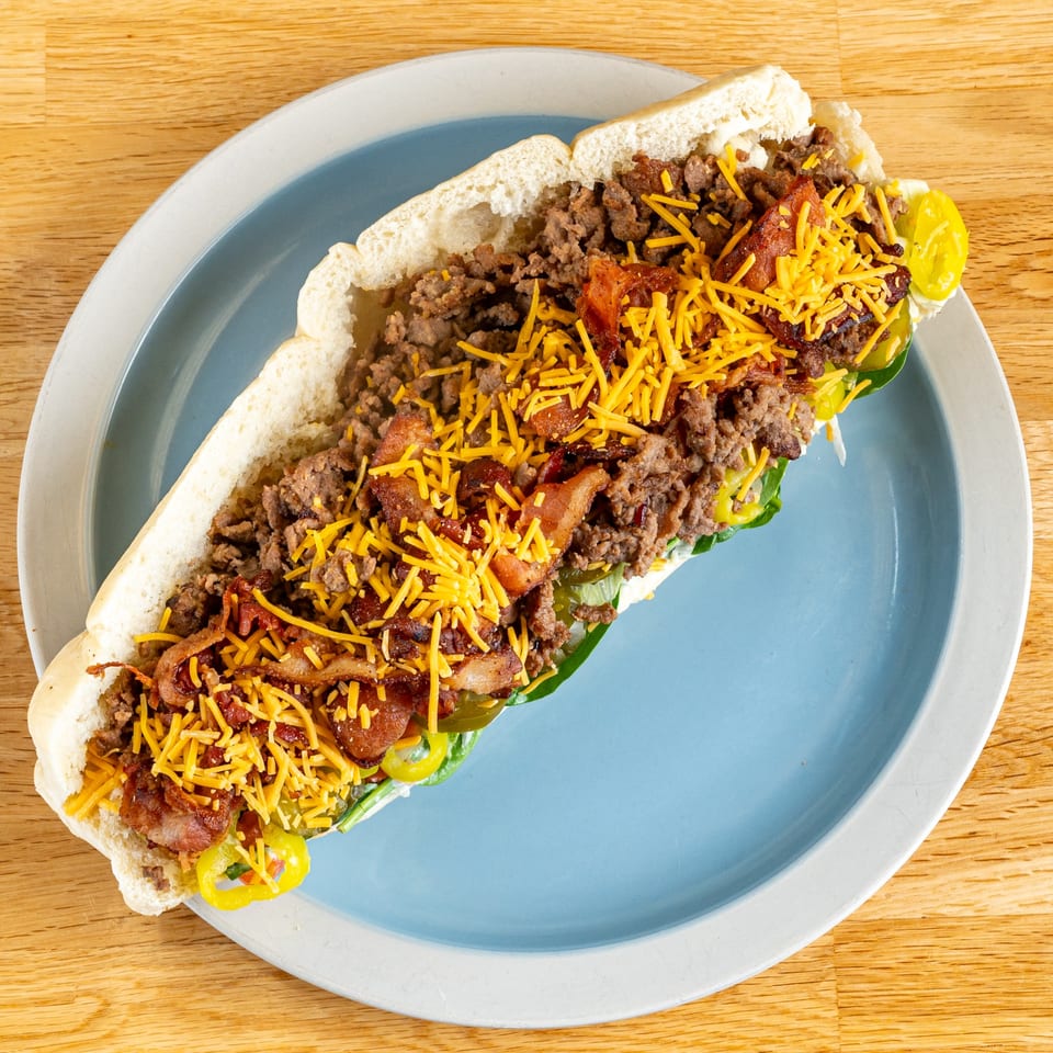 A loaded cheesesteak sub with ground beef, bacon, shredded cheese, and peppers on a blue plate.