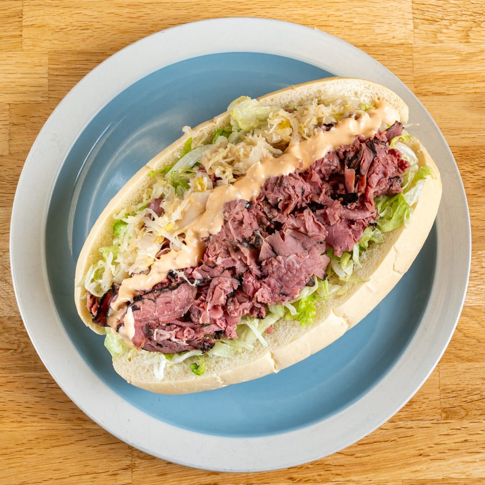 A Subs On The Run pastrami sub with sauerkraut, lettuce, and sauce on a blue plate.