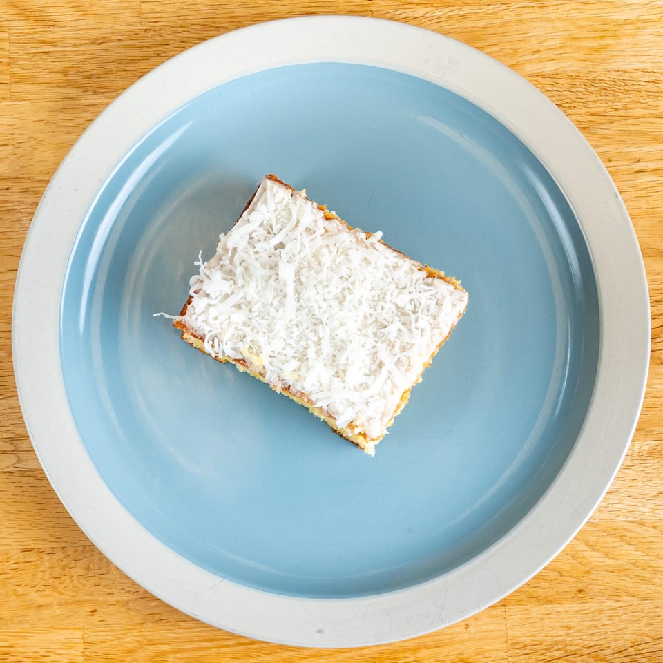 A square slice of cake with white frosting and shredded coconut on a blue and white plate.